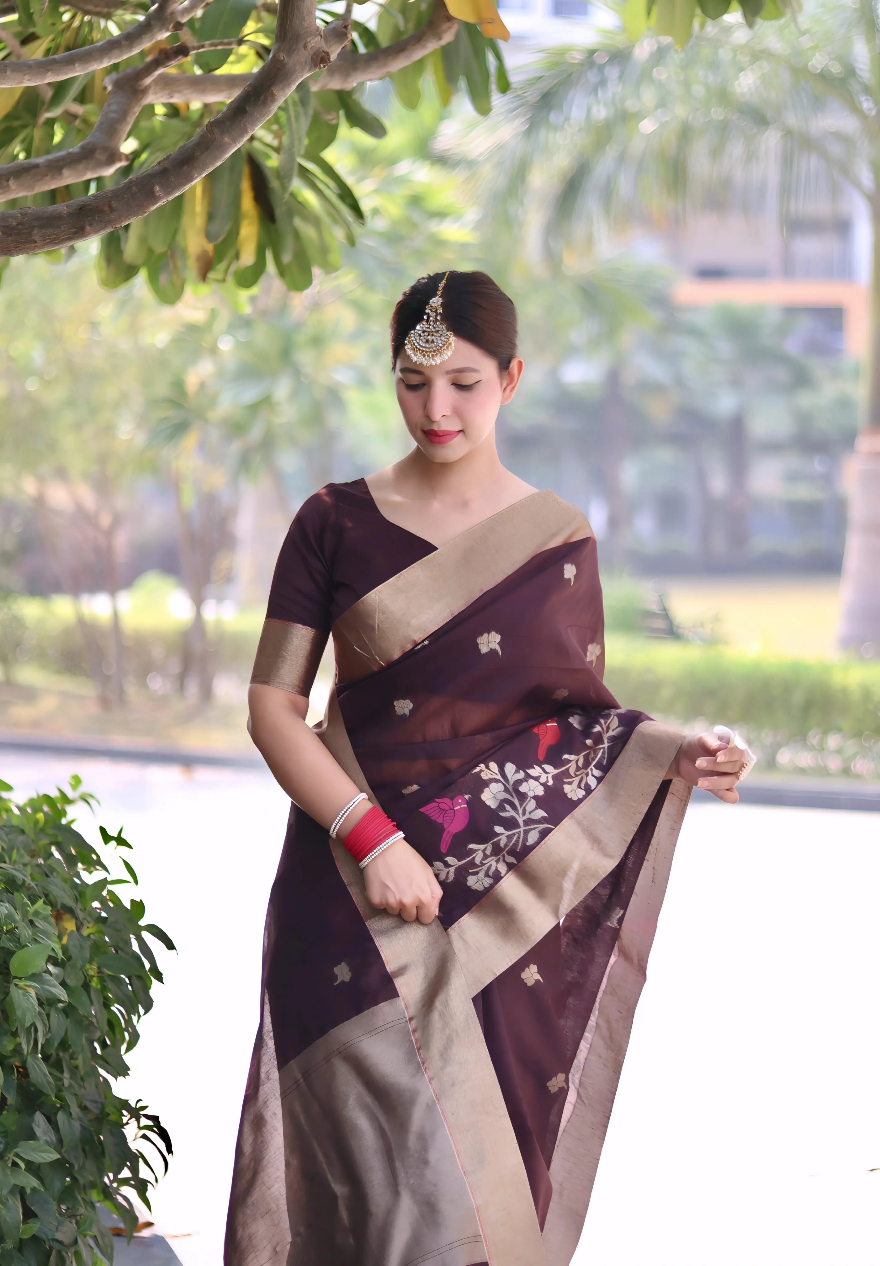 A woman models an elegant Deep Plum Linen Silk Saree. The saree features delicate woven floral and bird motifs and a wide, contrasting pale gold Zari border. She wears a matching plum blouse, contrasting red and white bangles, and a traditional gold maang tikka, posing outdoors.